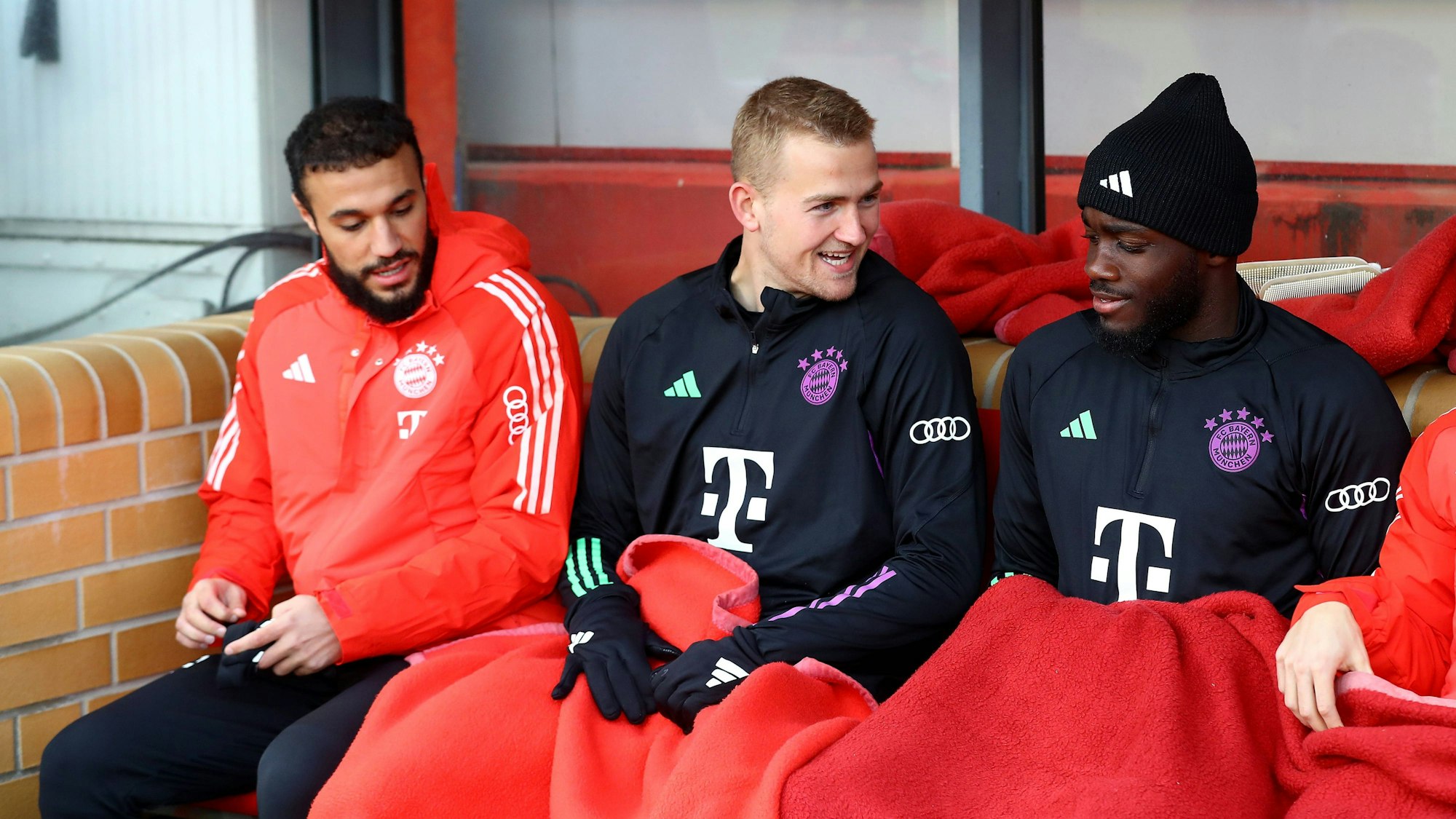Noussair Mazraoui (l.), Matthijs de Ligt (M.) und Dayot Upamecano sitzen auf der Ersatzbank bei einem Spiel des FC Bayern.
