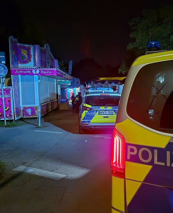 An einer Kirmes stehen abends zwei Streifenwagen, davor mehrere Polizeikräfte.