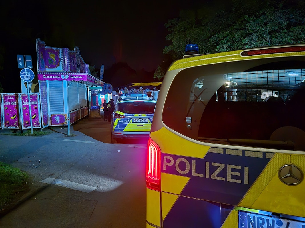 An einer Kirmes stehen abends zwei Streifenwagen, davor mehrere Polizeikräfte.