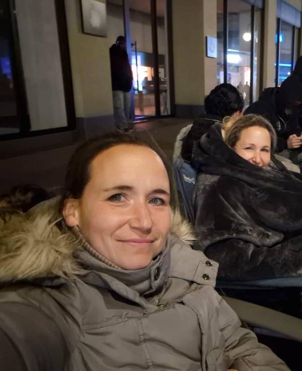 Melanie und ihre Schwester sitzen in Jacken und Decken verpackt auf Stühlen in einer Reihe von Menschen.
