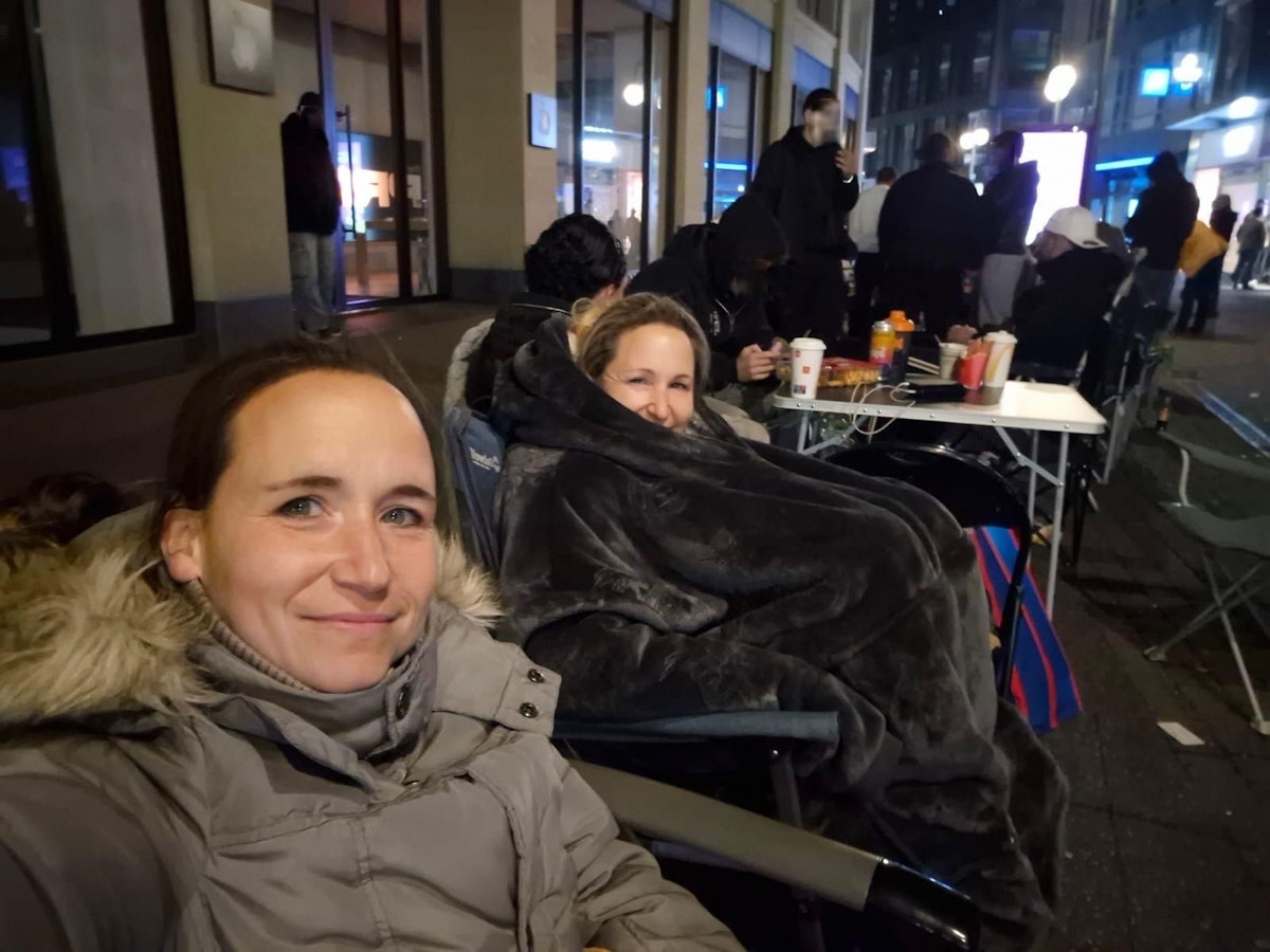 Melanie und ihre Schwester sitzen in Jacken und Decken verpackt auf Stühlen in einer Reihe von Menschen.