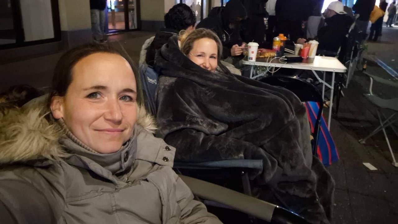 Melanie und ihre Schwester sitzen in Jacken und Decken verpackt auf Stühlen in einer Reihe von Menschen.