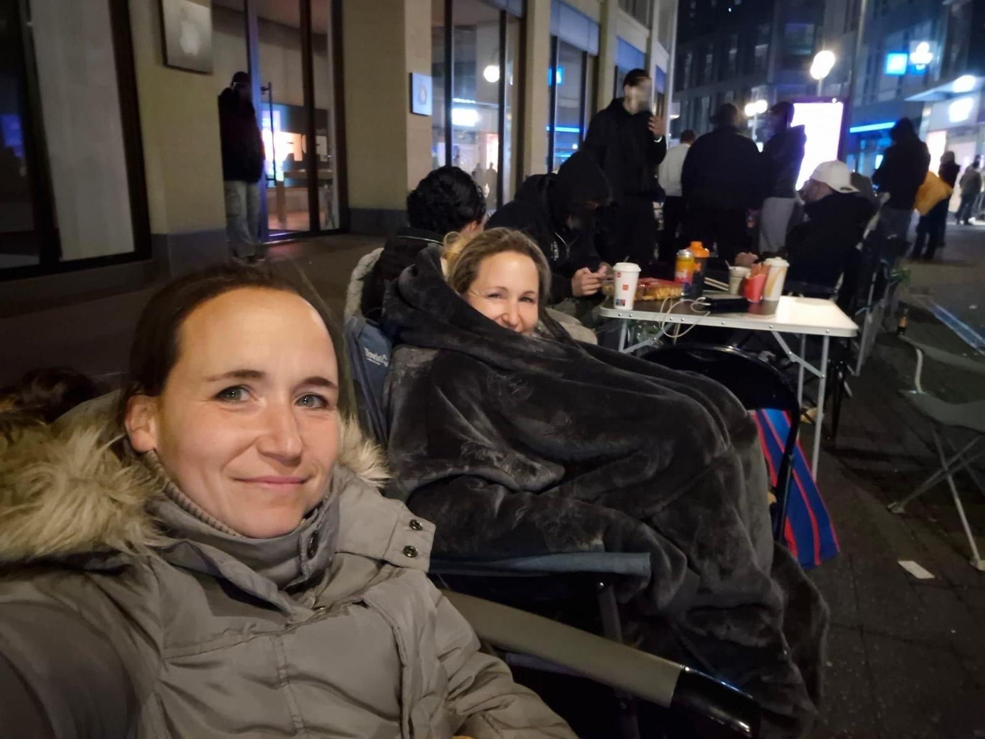 Melanie und ihre Schwester sitzen in Jacken und Decken verpackt auf Stühlen in einer Reihe von Menschen.