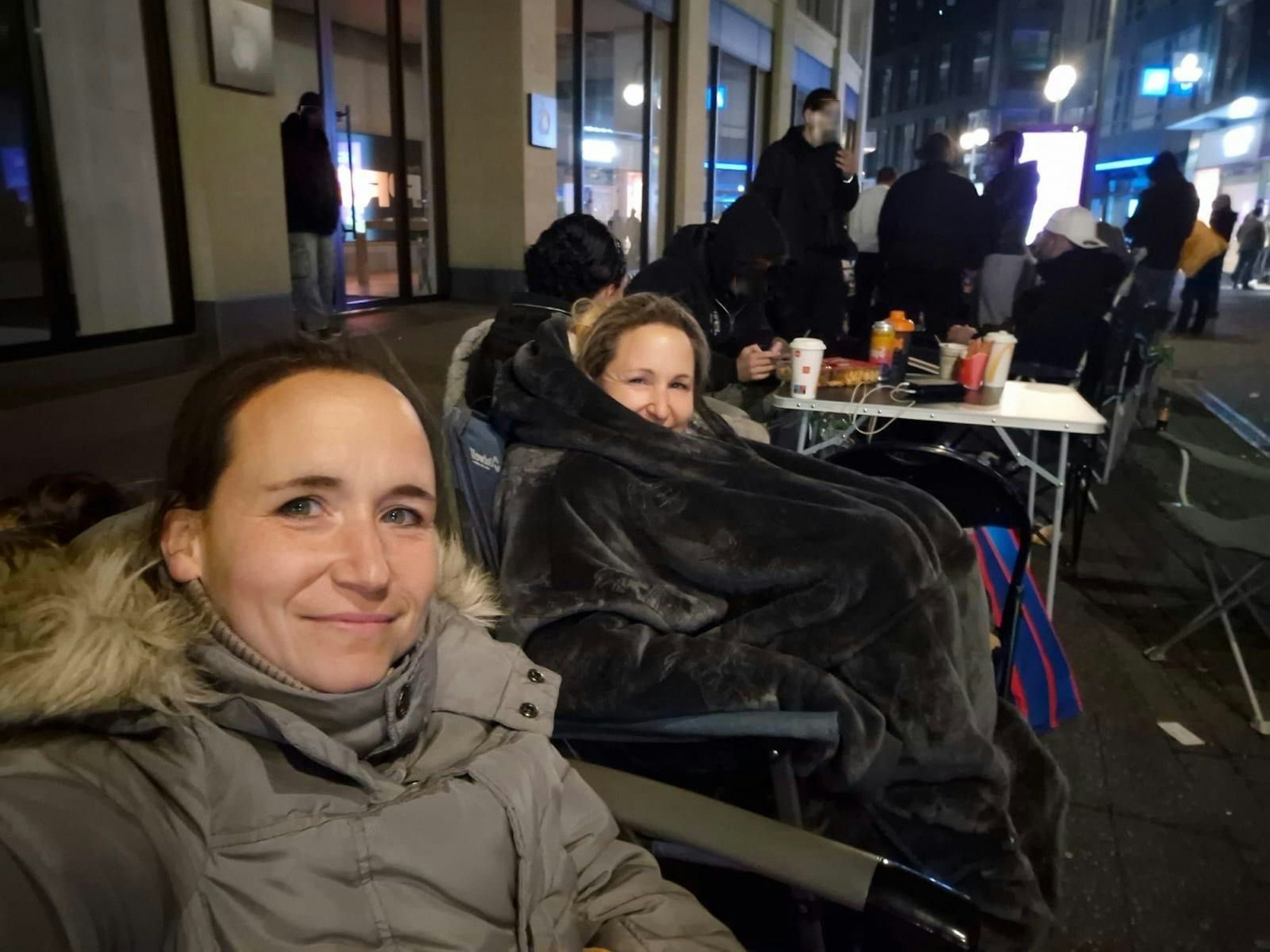Melanie und ihre Schwester sitzen in Jacken und Decken verpackt auf Stühlen in einer Reihe von Menschen.