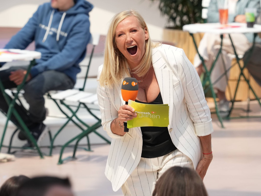 Andrea „Kiwi“ Kiewel lacht im ZDF Fernsehgarten, dabei hält sie Mikro und einen Zettel in der Hand.