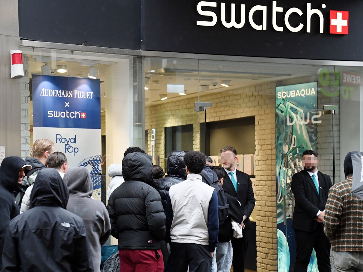 Menschen warten in einer Schlange vor dem Swatch-Store, vor dessen Tür stehen zwei Security-Mitarbeiter.