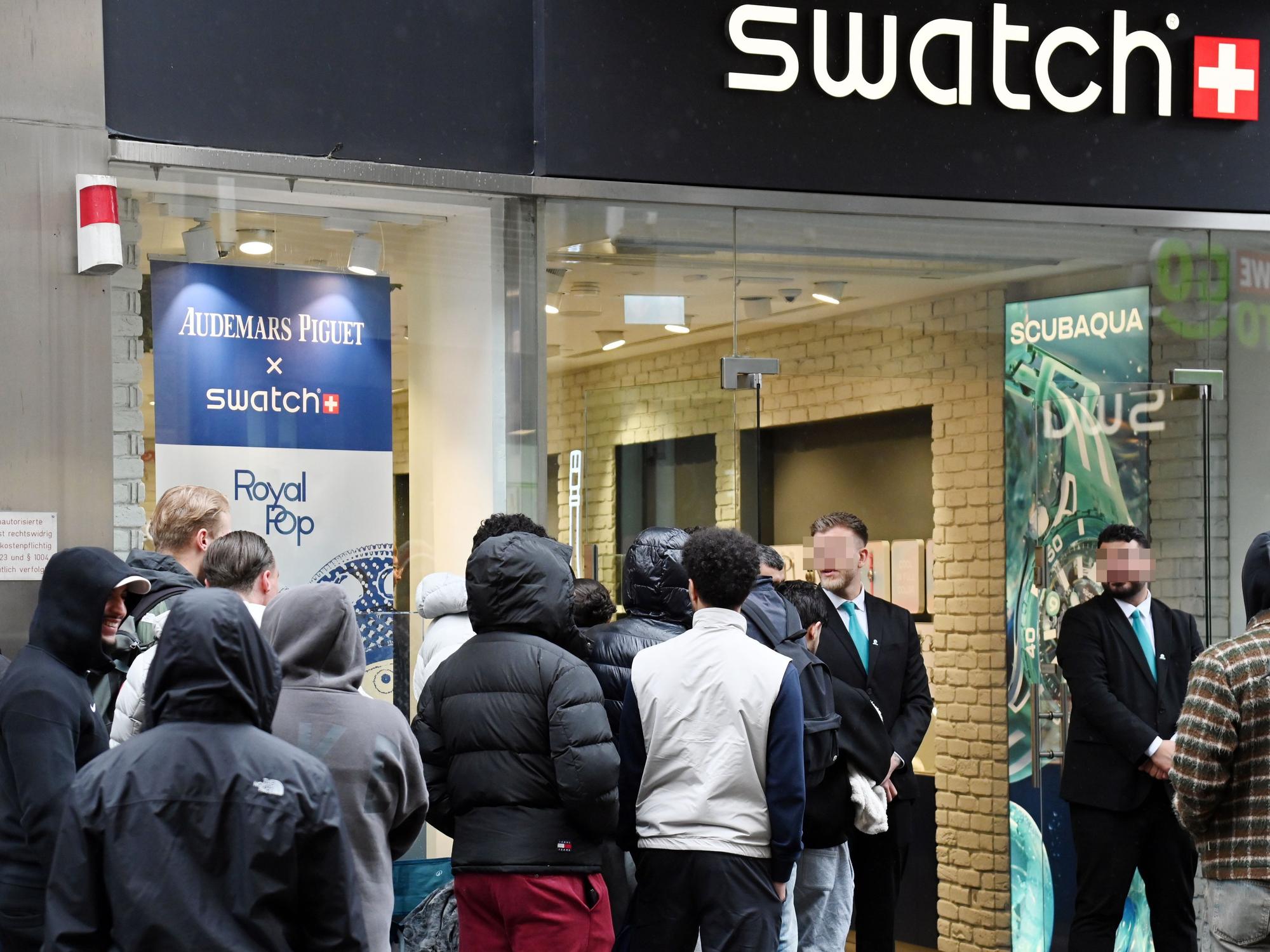 Menschen warten in einer Schlange vor dem Swatch-Store, vor dessen Tür stehen zwei Security-Mitarbeiter.