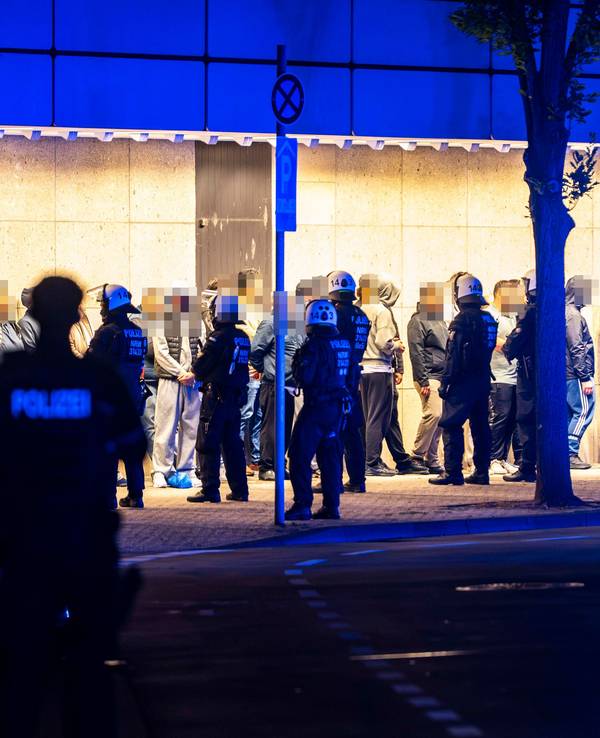 Menschen stehen aufgereiht vor einer Wand, vor ihnen stehen Polizisten.