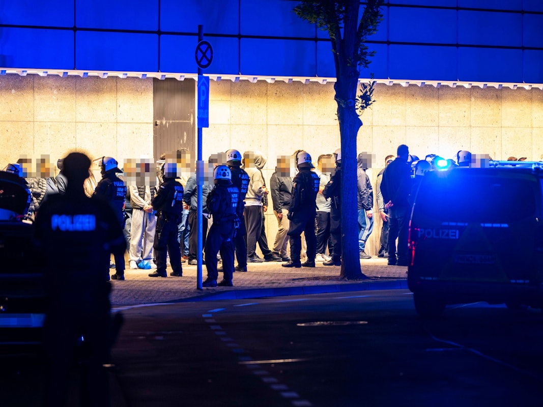 Menschen stehen aufgereiht vor einer Wand, vor ihnen stehen Polizisten.
