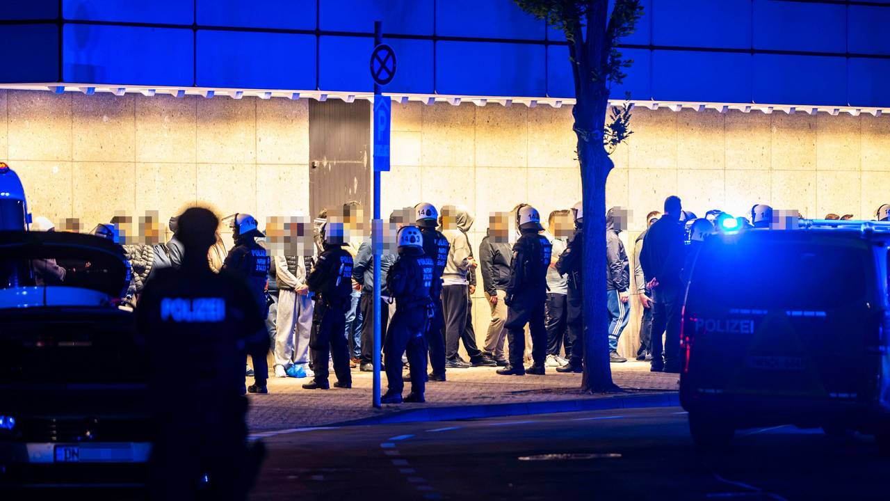 Menschen stehen aufgereiht vor einer Wand, vor ihnen stehen Polizisten.