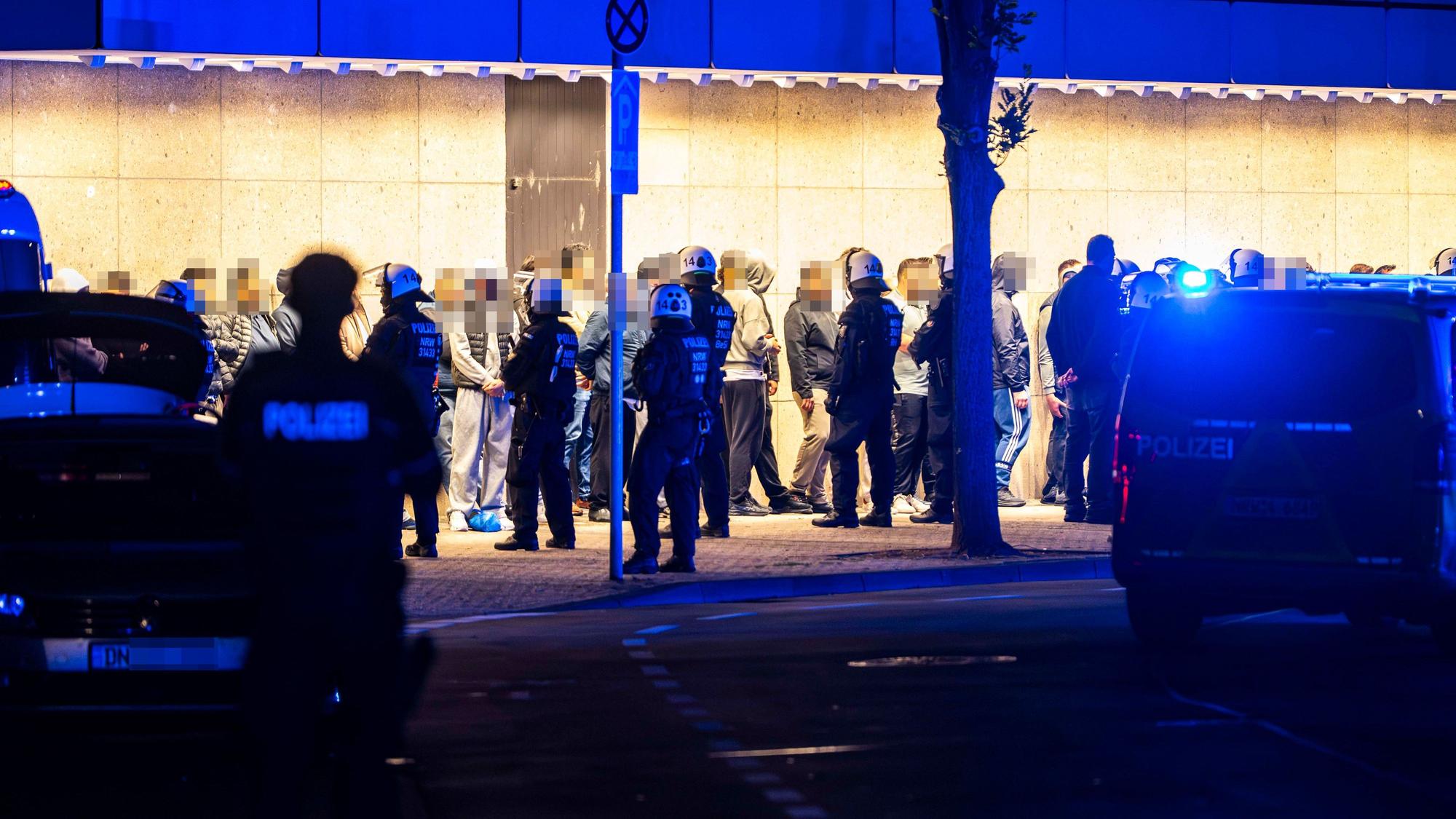Menschen stehen aufgereiht vor einer Wand, vor ihnen stehen Polizisten.