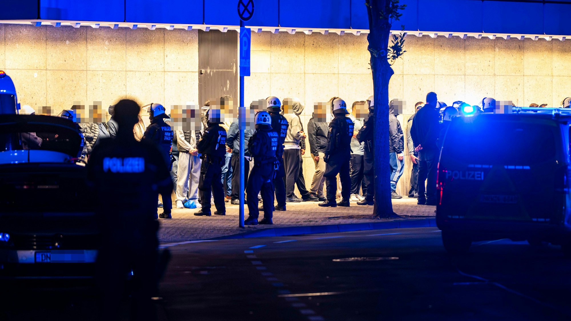 Menschen stehen aufgereiht vor einer Wand, vor ihnen stehen Polizisten.