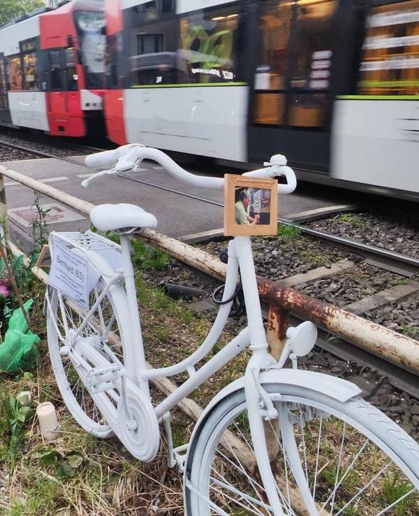 Ein weiß lackiertes Fahrrad steht an einem Bahnübergang während ein Zug vorbeirauscht.