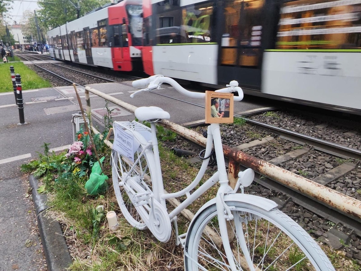 Ein weiß lackiertes Fahrrad steht an einem Bahnübergang während ein Zug vorbeirauscht.