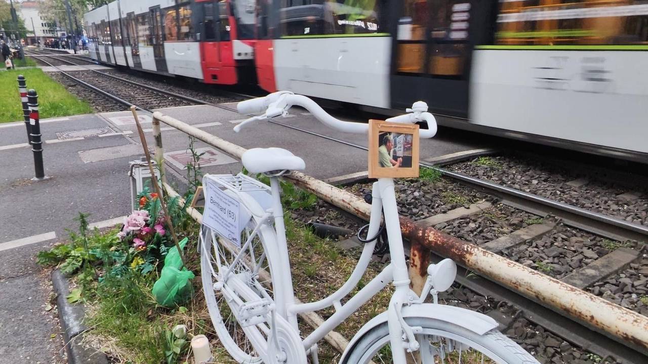 Ein weiß lackiertes Fahrrad steht an einem Bahnübergang während ein Zug vorbeirauscht.
