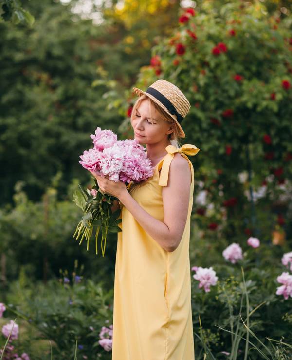 Eine Frau in einem Sommerkleid hält in Gedanken versunken einen Strauß Pfingstrosen vor sich