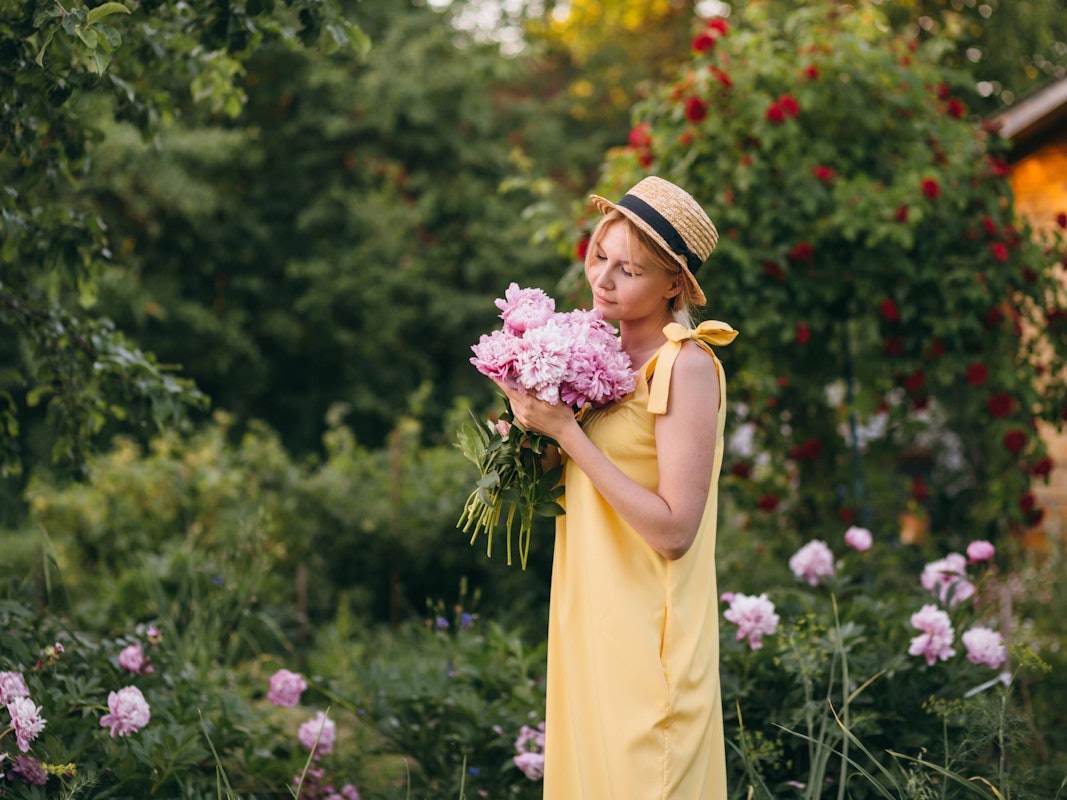 Eine Frau in einem Sommerkleid hält in Gedanken versunken einen Strauß Pfingstrosen vor sich