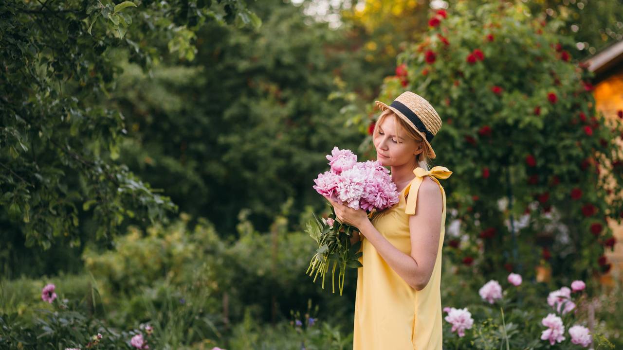 Eine Frau in einem Sommerkleid hält in Gedanken versunken einen Strauß Pfingstrosen vor sich