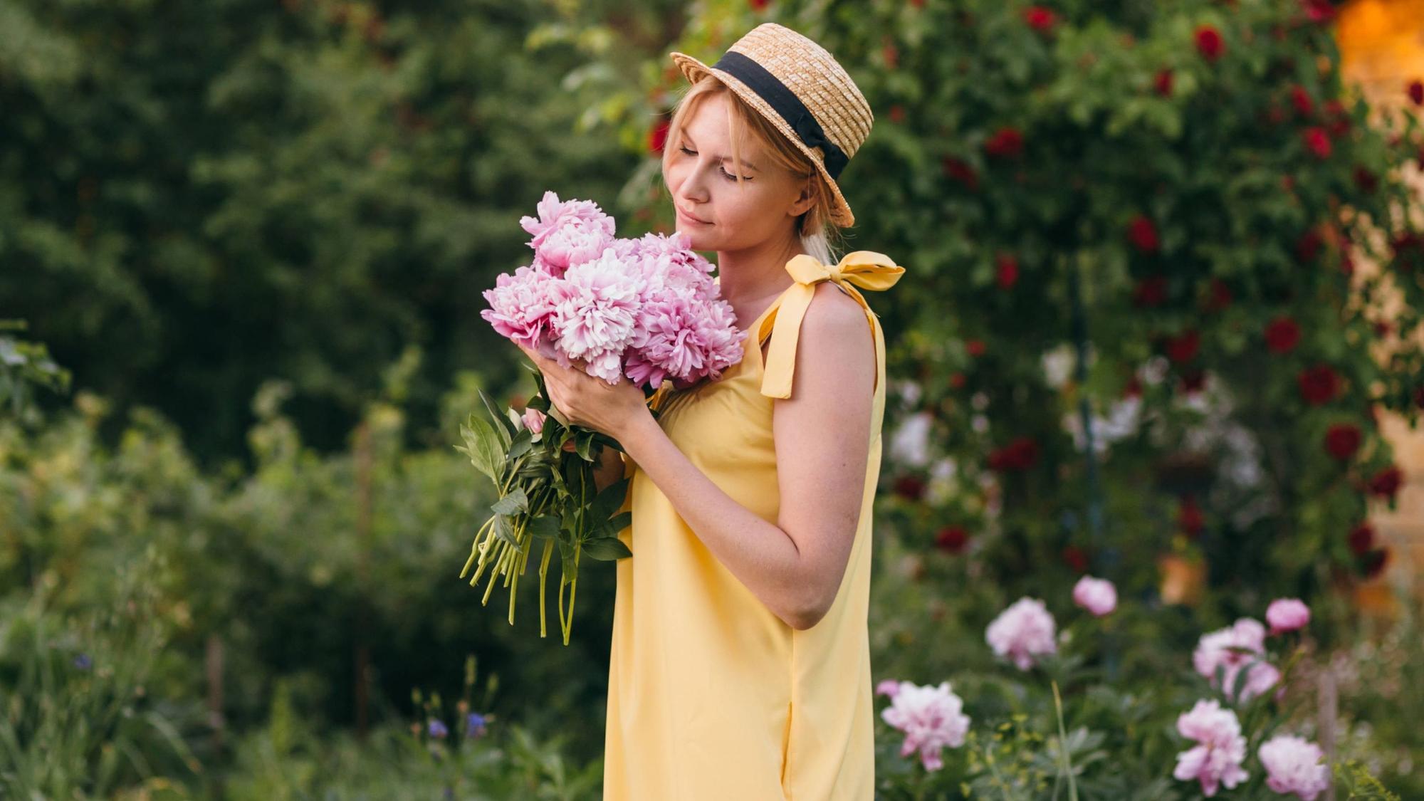 Eine Frau in einem Sommerkleid hält in Gedanken versunken einen Strauß Pfingstrosen vor sich