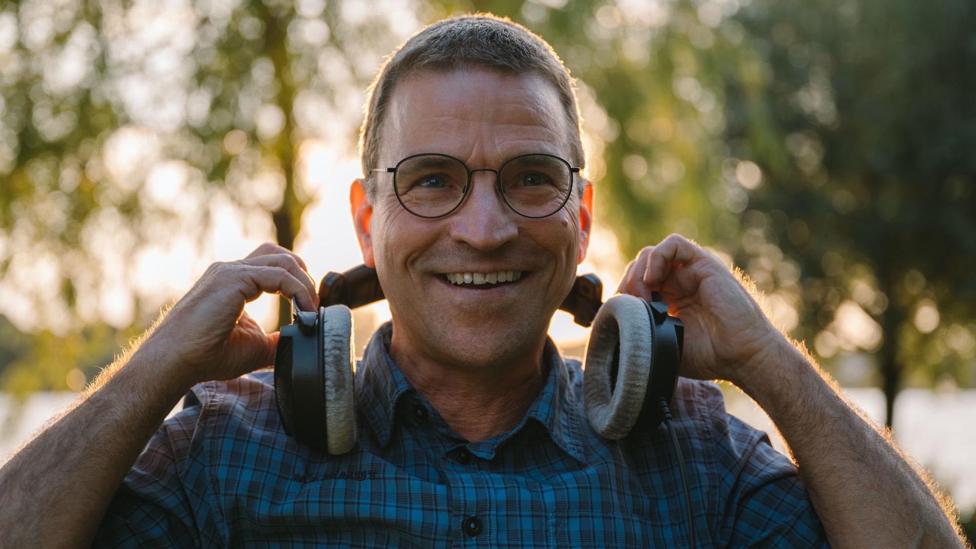 Ein älterer Herr mit Brille lacht in die Kamera und nimmt sich Kopfhörer vom Kopf