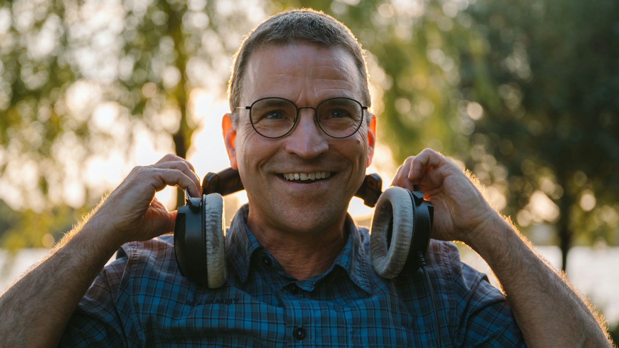 Ein älterer Herr mit Brille lacht in die Kamera und nimmt sich Kopfhörer vom Kopf