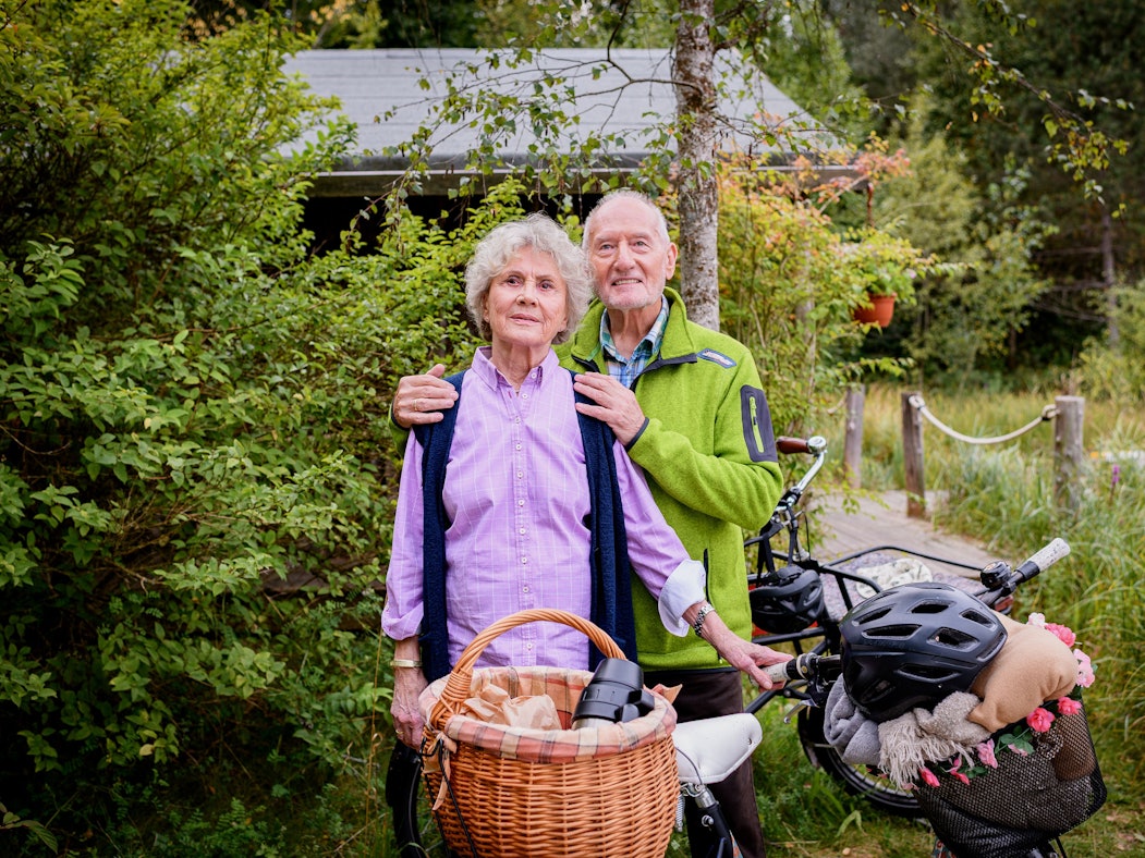 Ein älteres Paar mit Fahrraädern und Funktionsklamotten steht zusammen, der Man umarmt die Frau sanft von hinten.