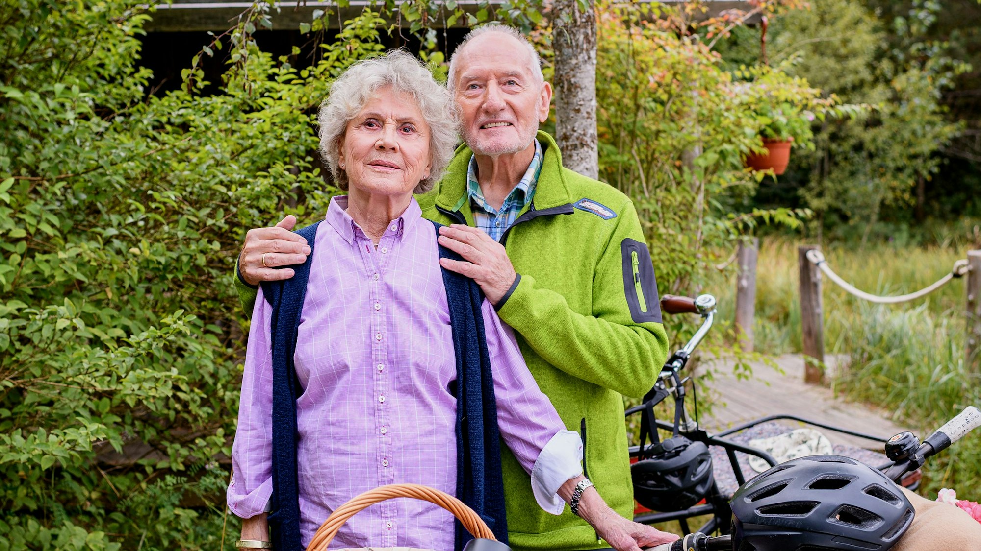 Ein älteres Paar mit Fahrraädern und Funktionsklamotten steht zusammen, der Man umarmt die Frau sanft von hinten.