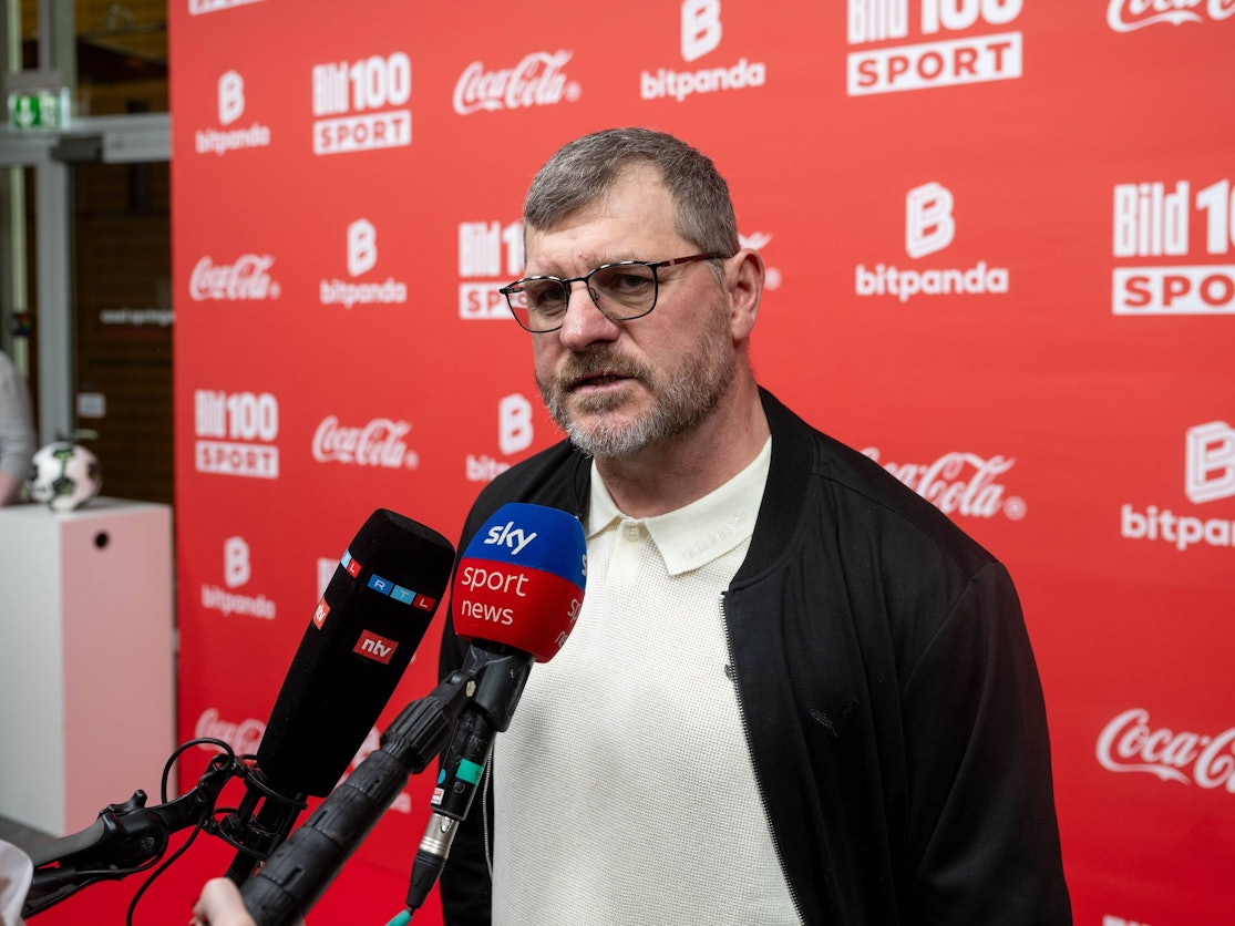 Fußballtrainer Steffen Baumgart steht vor mehreren Mikrofonen bei einer Veranstaltung.