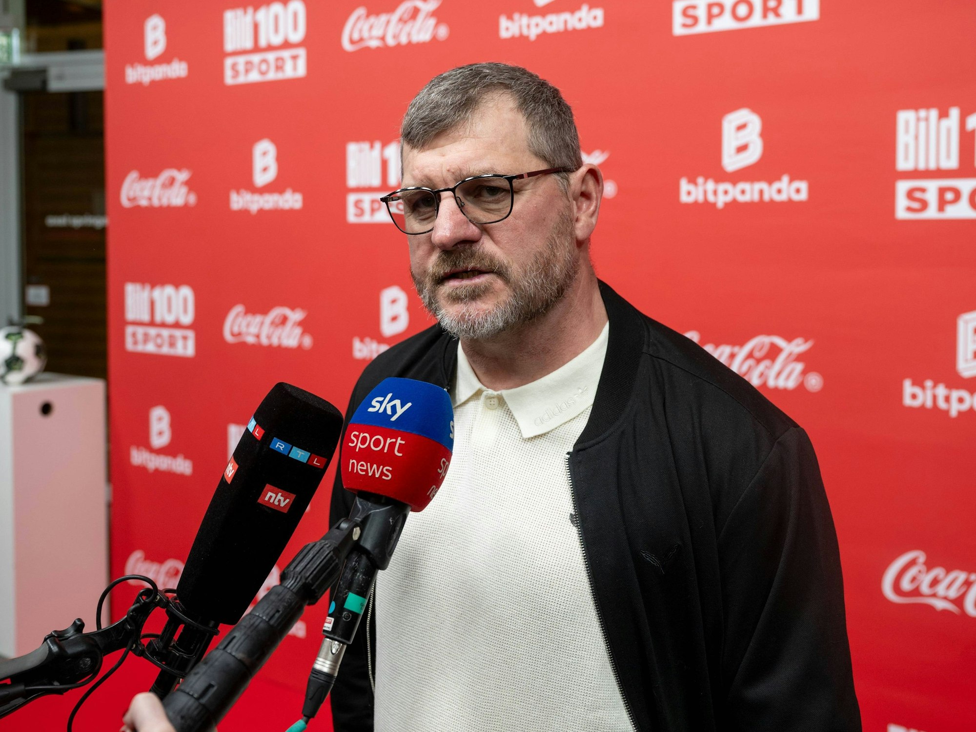 Fußballtrainer Steffen Baumgart steht vor mehreren Mikrofonen bei einer Veranstaltung.