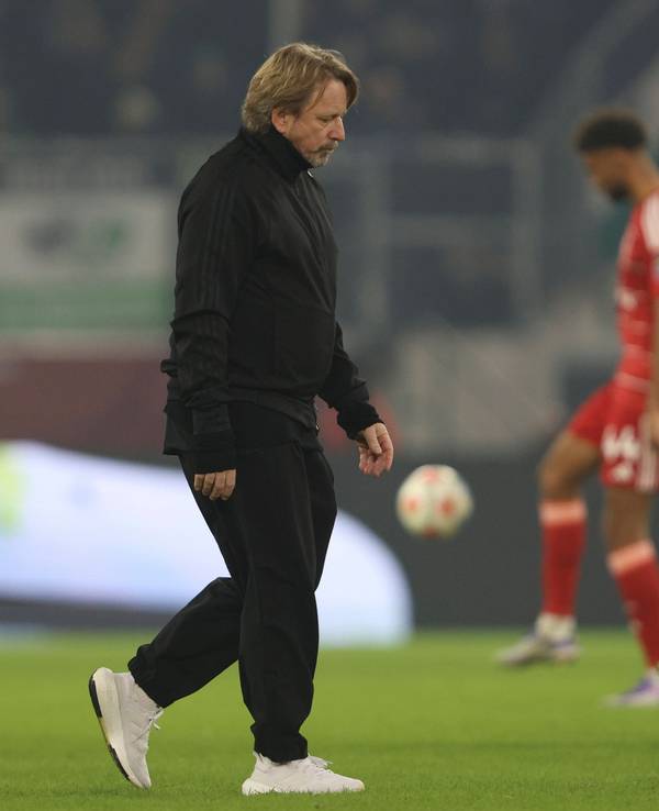Sven Mislintat geht mit enttäuschtem Blick über den Rasen. Im Hintergrund sieht man zwei Spieler der Fortuna, die sich aufwärmen.