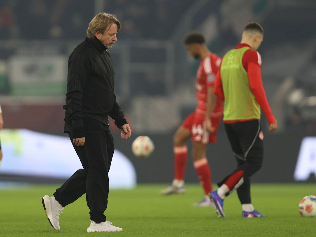 Sven Mislintat geht mit enttäuschtem Blick über den Rasen. Im Hintergrund sieht man zwei Spieler der Fortuna, die sich aufwärmen.