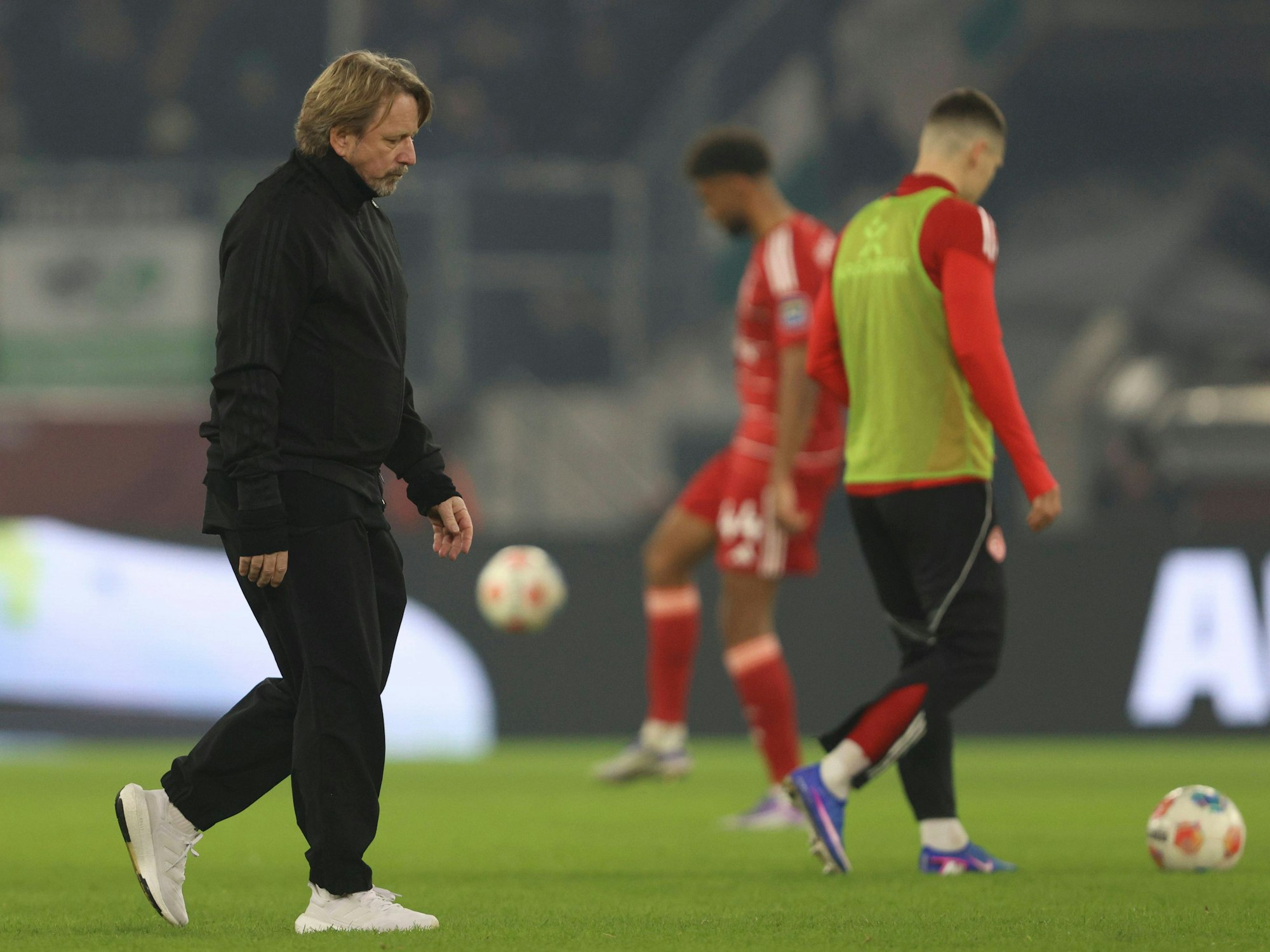 Sven Mislintat geht mit enttäuschtem Blick über den Rasen. Im Hintergrund sieht man zwei Spieler der Fortuna, die sich aufwärmen.