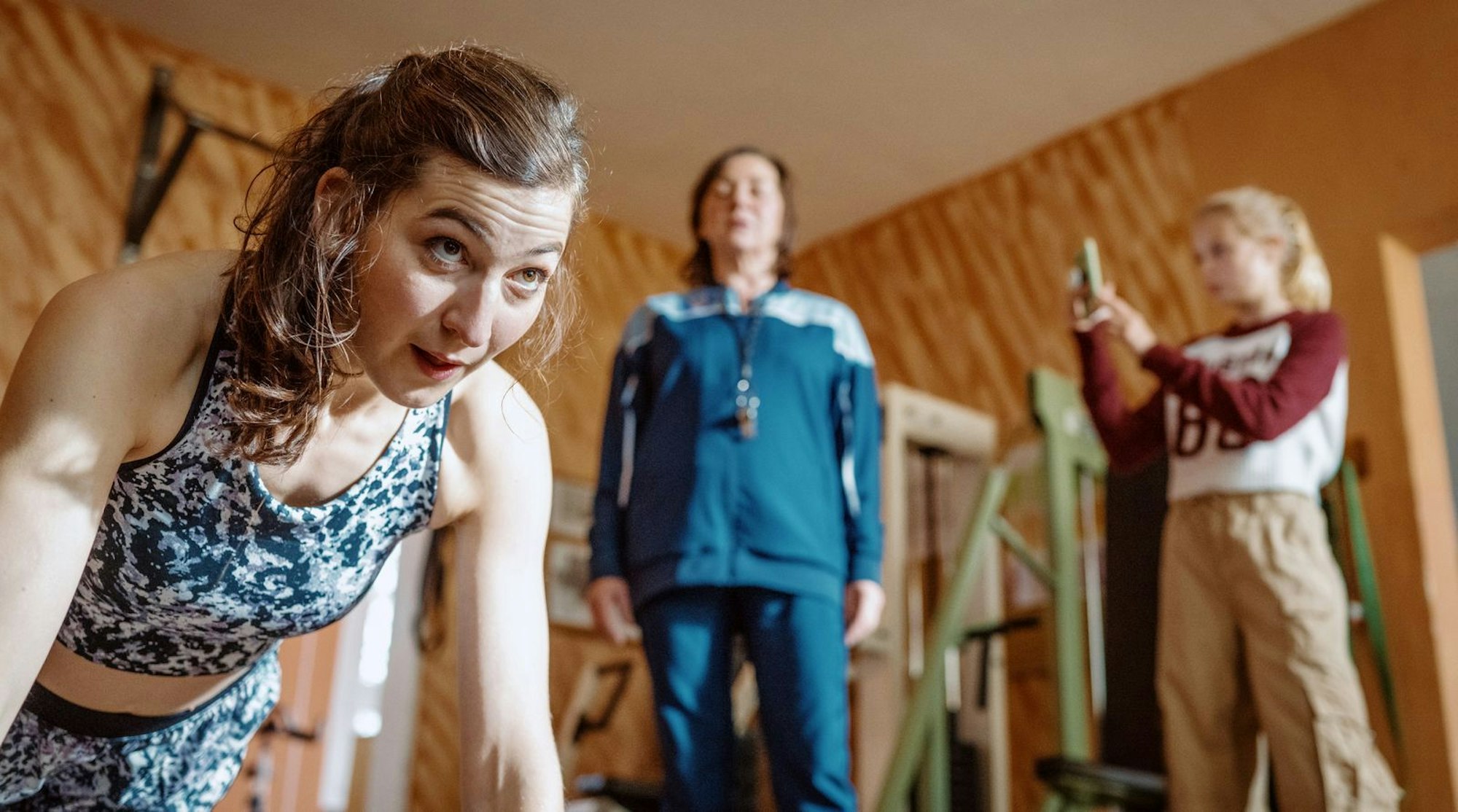 „Fit in Gerlau“ soll über Social Media beworben werden: Enkelin Emma (Selma Suckfüll, rechts) filmt Mitarbeiterin Katleen (Ann-Sophie Schindler, links). Oma Christa (Steffi Kühnert) gibt die Einpeitscherin. (Bild: © ARD Degeto Film/Adrian Gross)