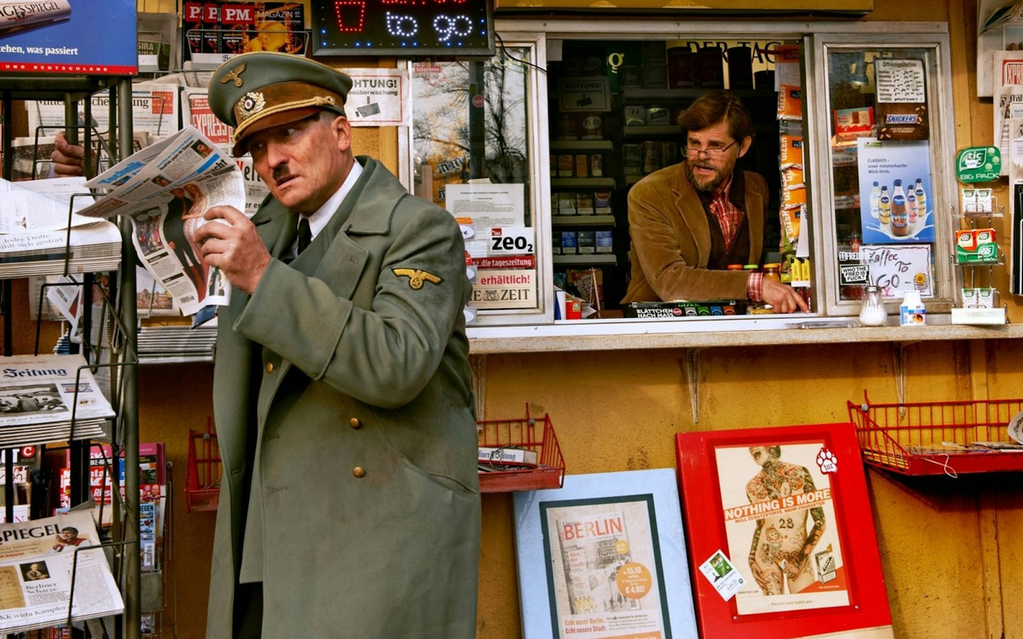 Der erste Weg für den neu erwachten Hitler (Oliver Masucci, links) zu einem Kiosk, an dem ihm seine Situation bewusst wird. Er lebt nun im Jahr 2014. Den „Krämer“ spielt Lars Rudolph. (Bild: 2015 Constantin Film Verleih GmbH)