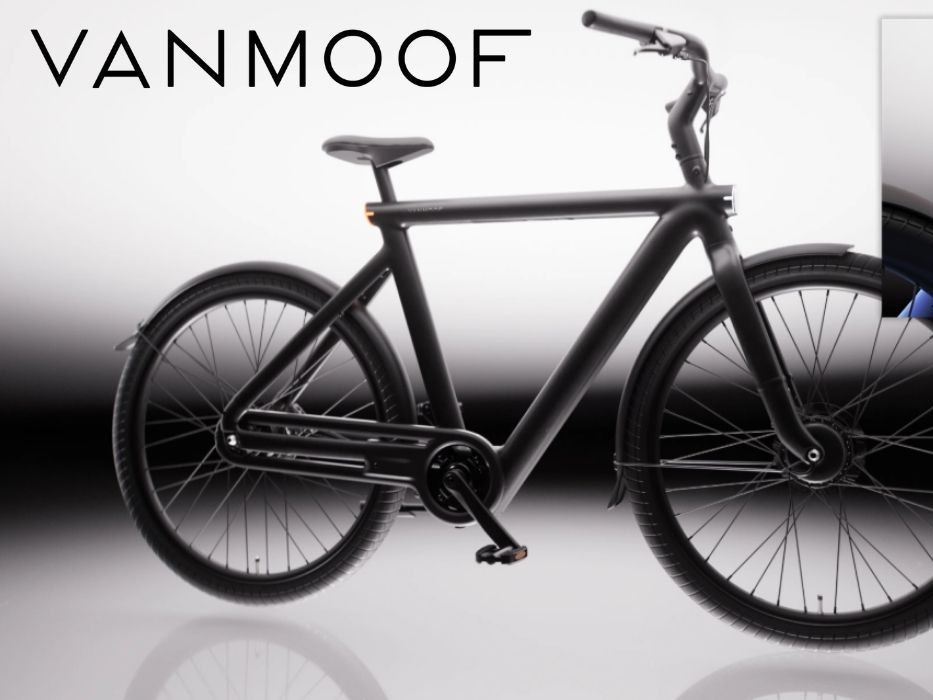 VanMoof S6 E-Bike in der Seitenansicht in der Farbe Drift Black vor hellem Hintergrund, mit Detailaufnahme des nahtlos in den Aluminiumrahmen integrierten Rücklichts in der Farbe Electric Blue – sichtbar auch das integrierte Frontlicht am Vorderrad.