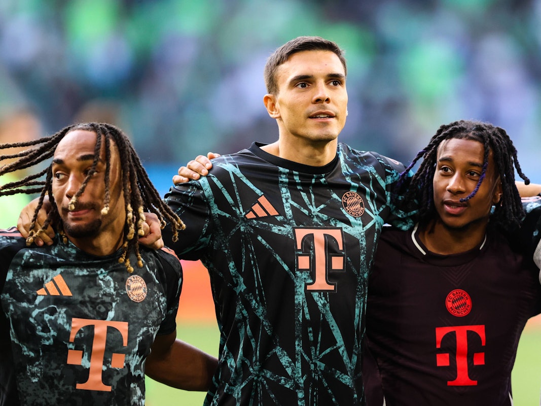 Sacha Boey (l.), Joao Palhinha (M.) und Michael Olise stehen Arm in Arm vor der Fankurve und feiern.