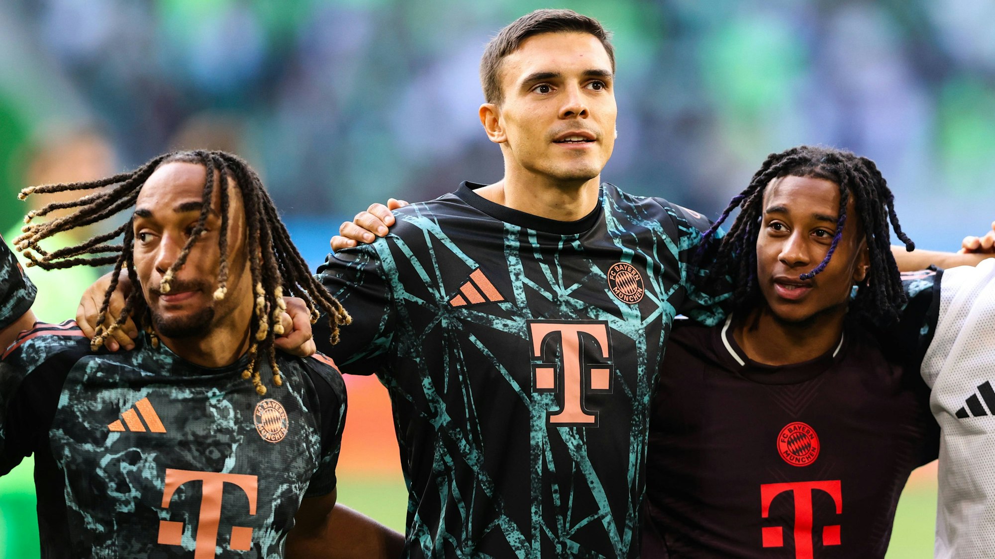 Sacha Boey (l.), Joao Palhinha (M.) und Michael Olise stehen Arm in Arm vor der Fankurve und feiern.