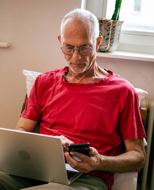 Ein Senior sitzt mit Laptop und Smartphone auf einem Sessel