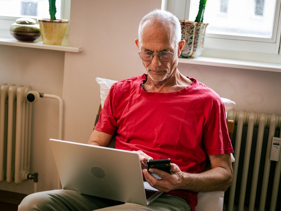 Ein Senior sitzt mit Laptop und Smartphone auf einem Sessel