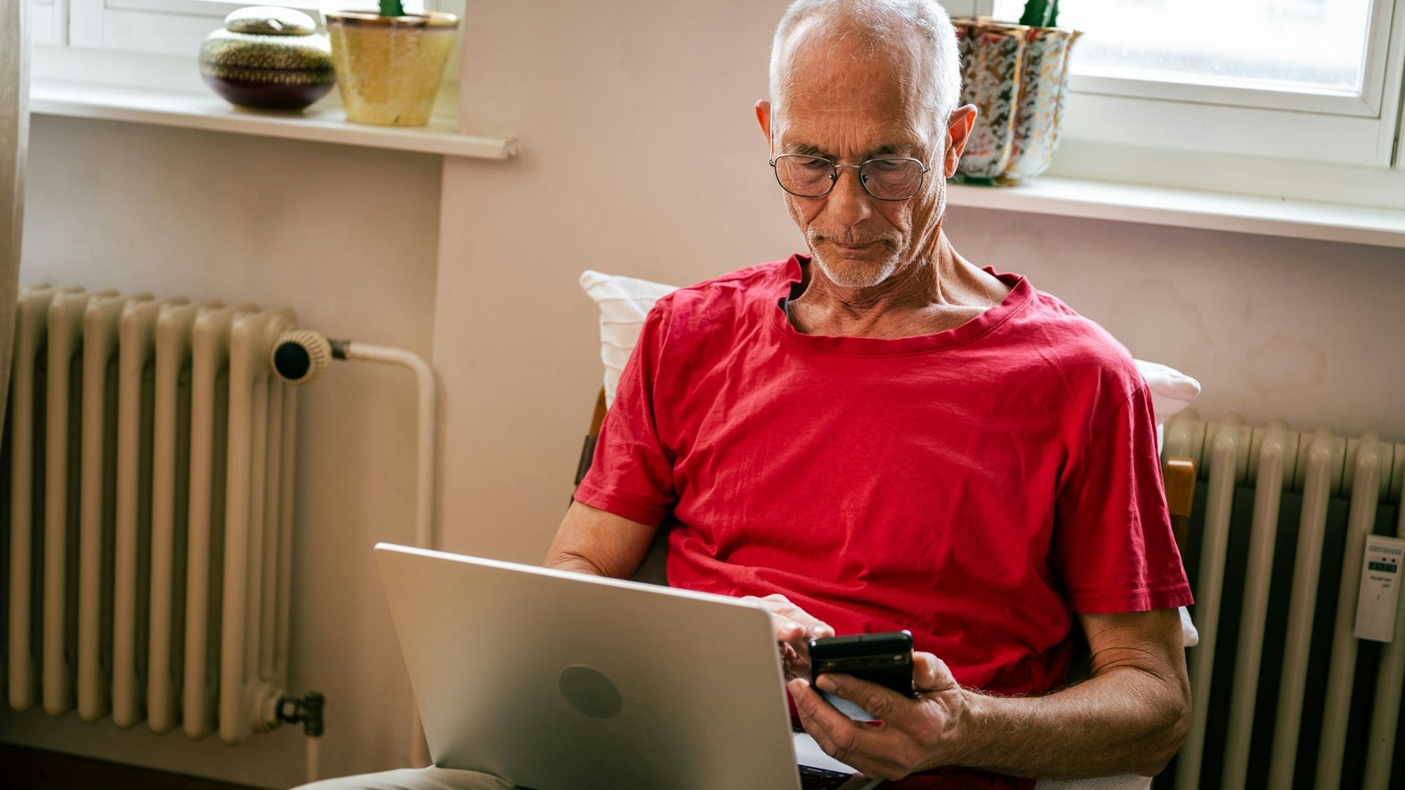 Ein Senior sitzt mit Laptop und Smartphone auf einem Sessel