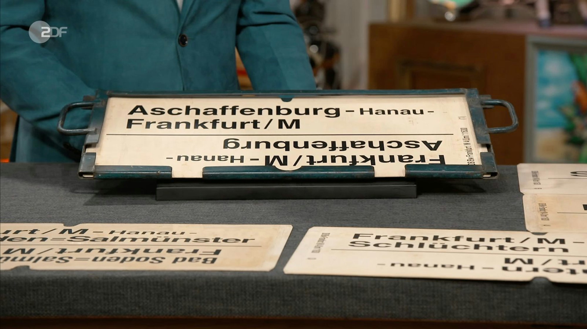 Als weitere Objekte der Sendung wurden fünf Zuglaufschilder samt Halterung aus dem regionalen Reiseverkehr der Deutschen Bahn von Detlev Kümmel in den 1970er-Jahren datiert. Gewünscht wurden 150 bis 200 Euro, Kümmel schätzte 100 bis 150 Euro und Händler Meyer zahlte 130 Euro. (Bild: ZDF)