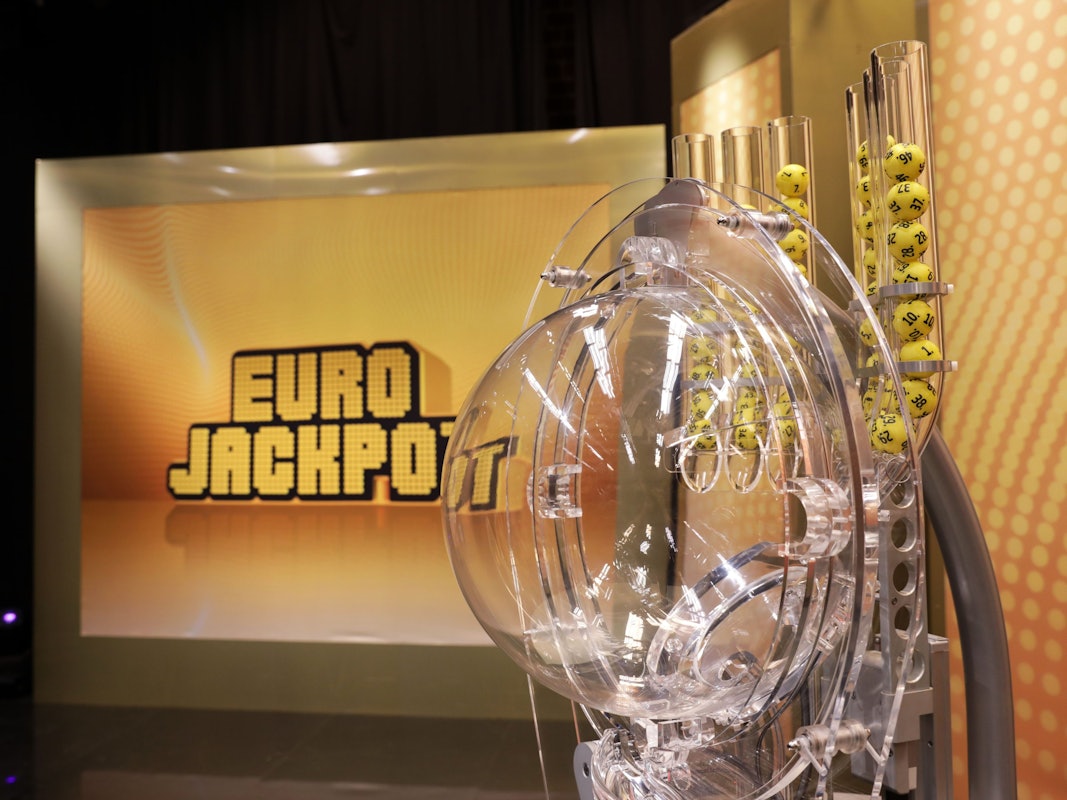 Das Bild zeigt die Ziehungsmaschine für die Eurojackpot-Lotterie. Im Hintergrund ist groß das goldene „EURO JACKPOT“-Logo auf einer beleuchteten Wand zu sehen.