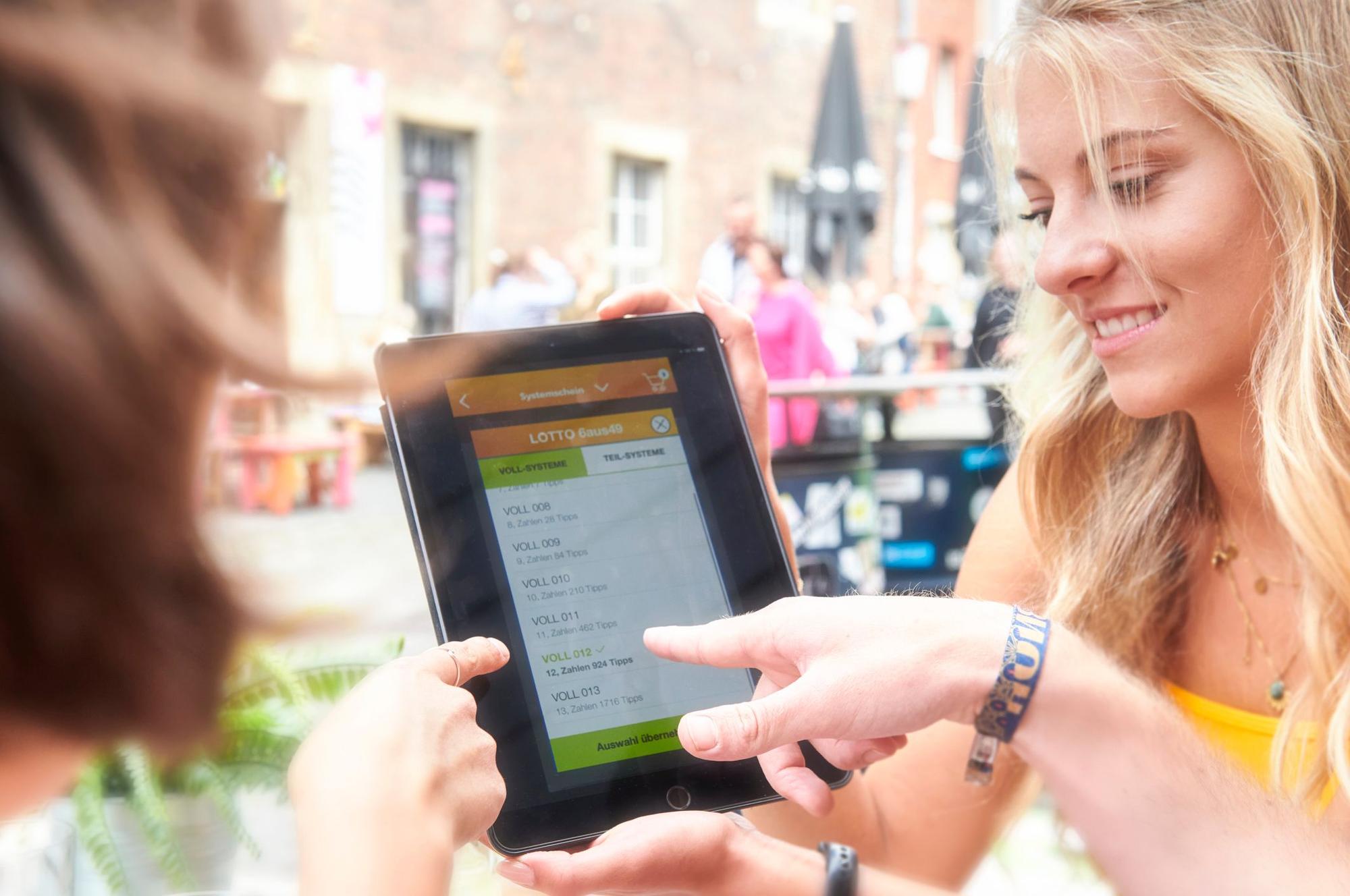 Zwei Personen schauen gemeinsam auf ein Tablet, auf dem die Auswahl für LOTTO 6aus49 zu sehen ist.