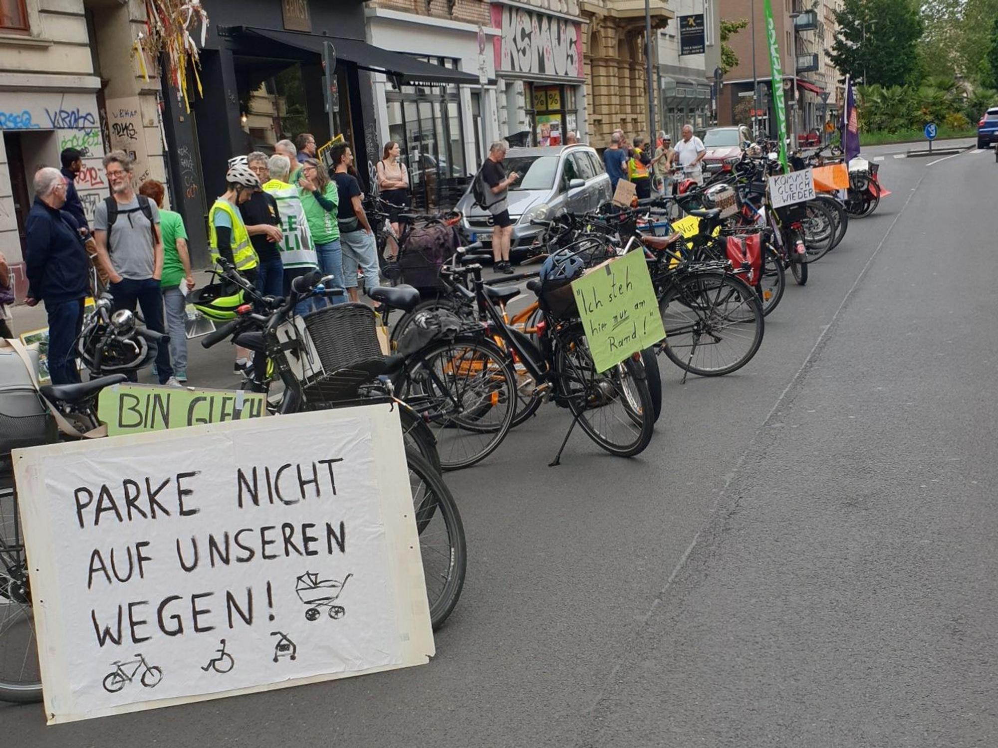 Fahrräder stehen auf der Fahrbahn, an ihnen hängen Plakate u. a. mit der Aufschrift: Parke nicht auf unseren Wegen!
