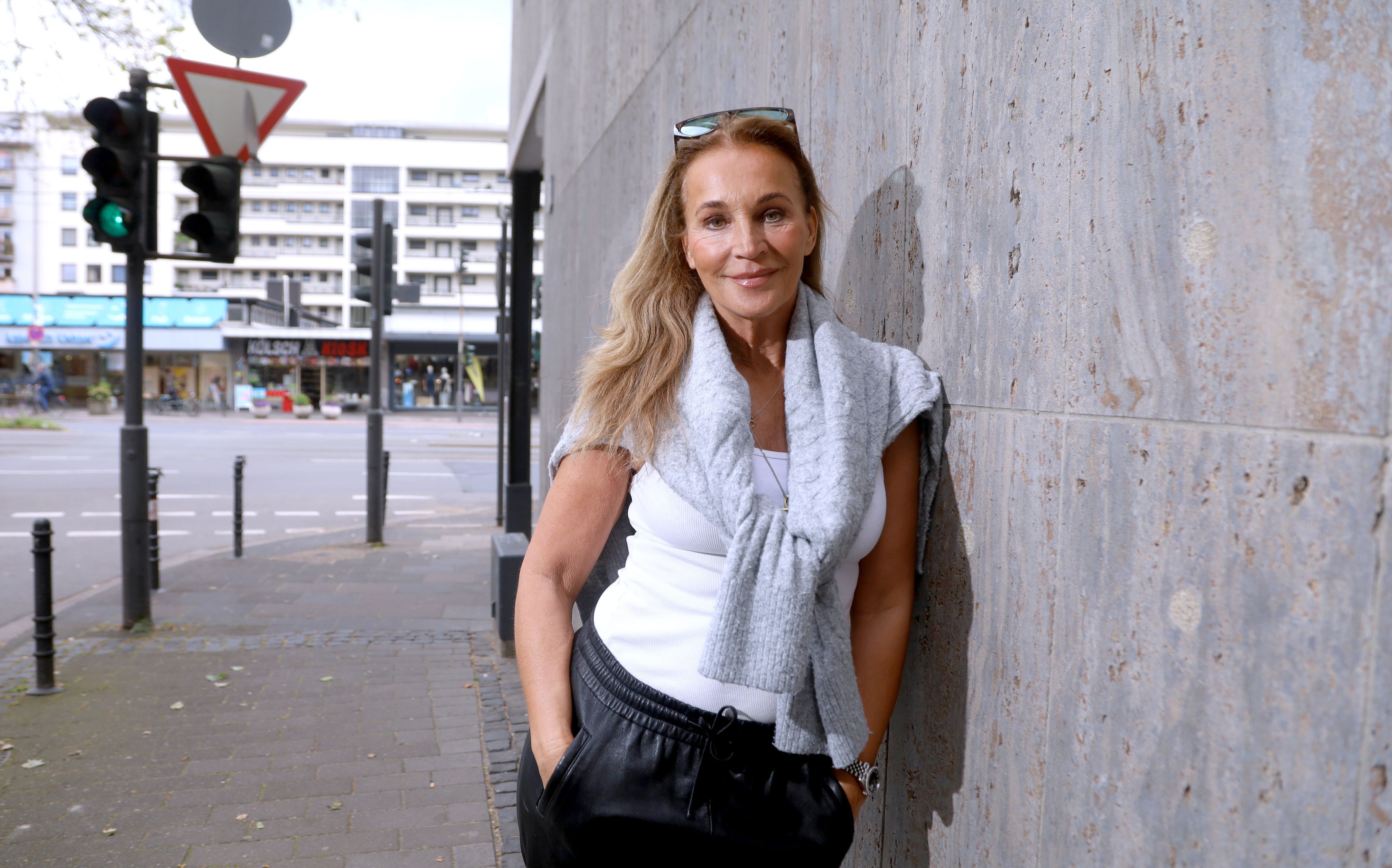Eine Frau in lockerer Sportkleidung mit Sonnenbrille im Haaar lehnt lässig an einer Mauer