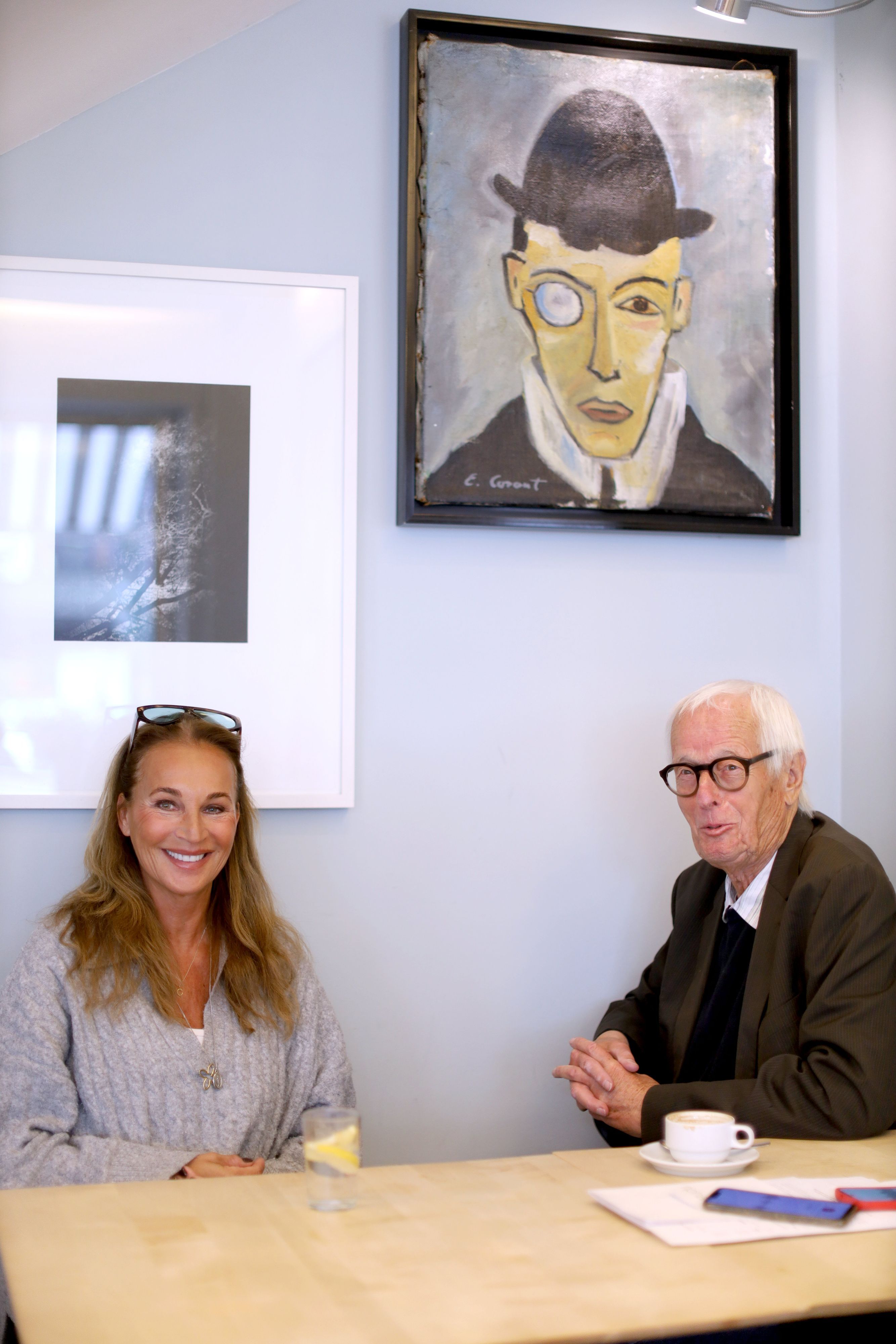 Eine Frau mit langen, brünetten Haaren und ein älterer Herr mit markanter Brille sitzen an einem Tisch und lachen freundlich in die Kamera