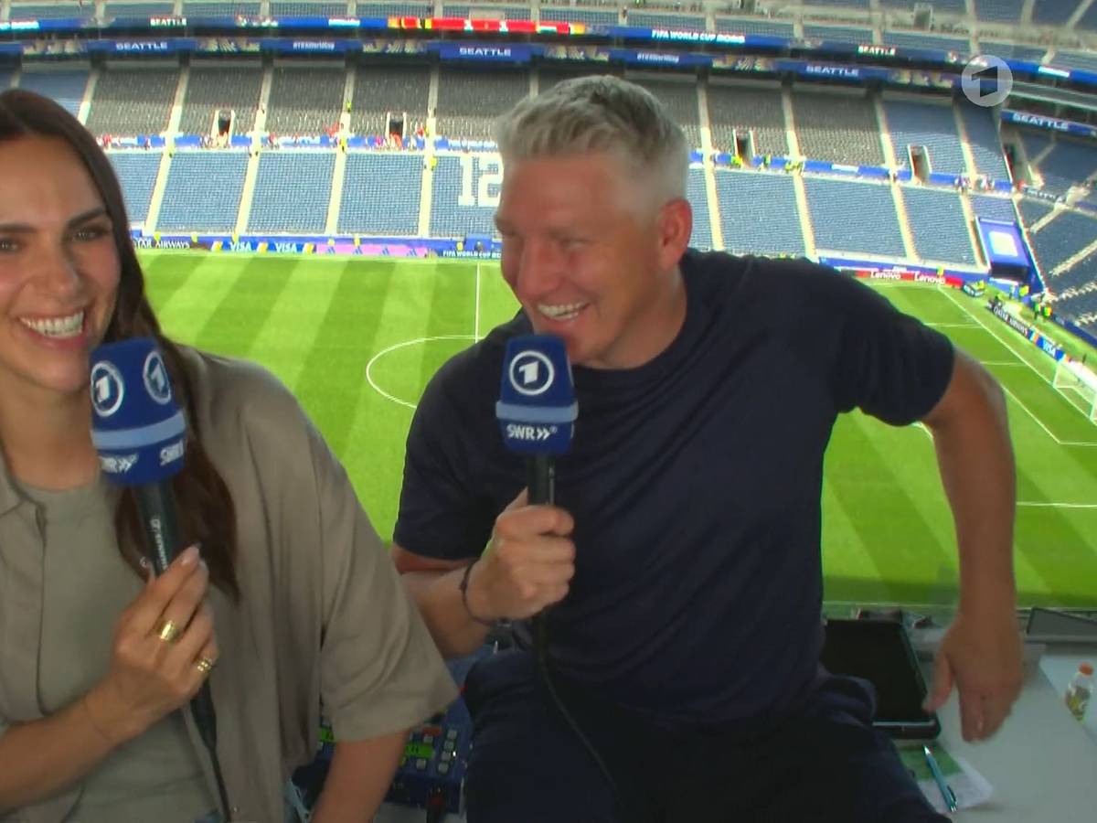 Esther Sedlaczek und Bastian Schweinsteiger sitzen nebeneinander im Stadion und lachen.