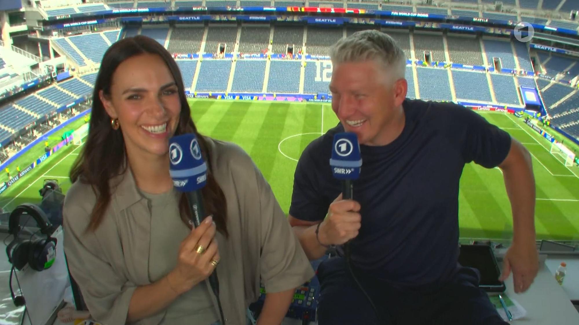 Esther Sedlaczek und Bastian Schweinsteiger sitzen nebeneinander im Stadion und lachen.