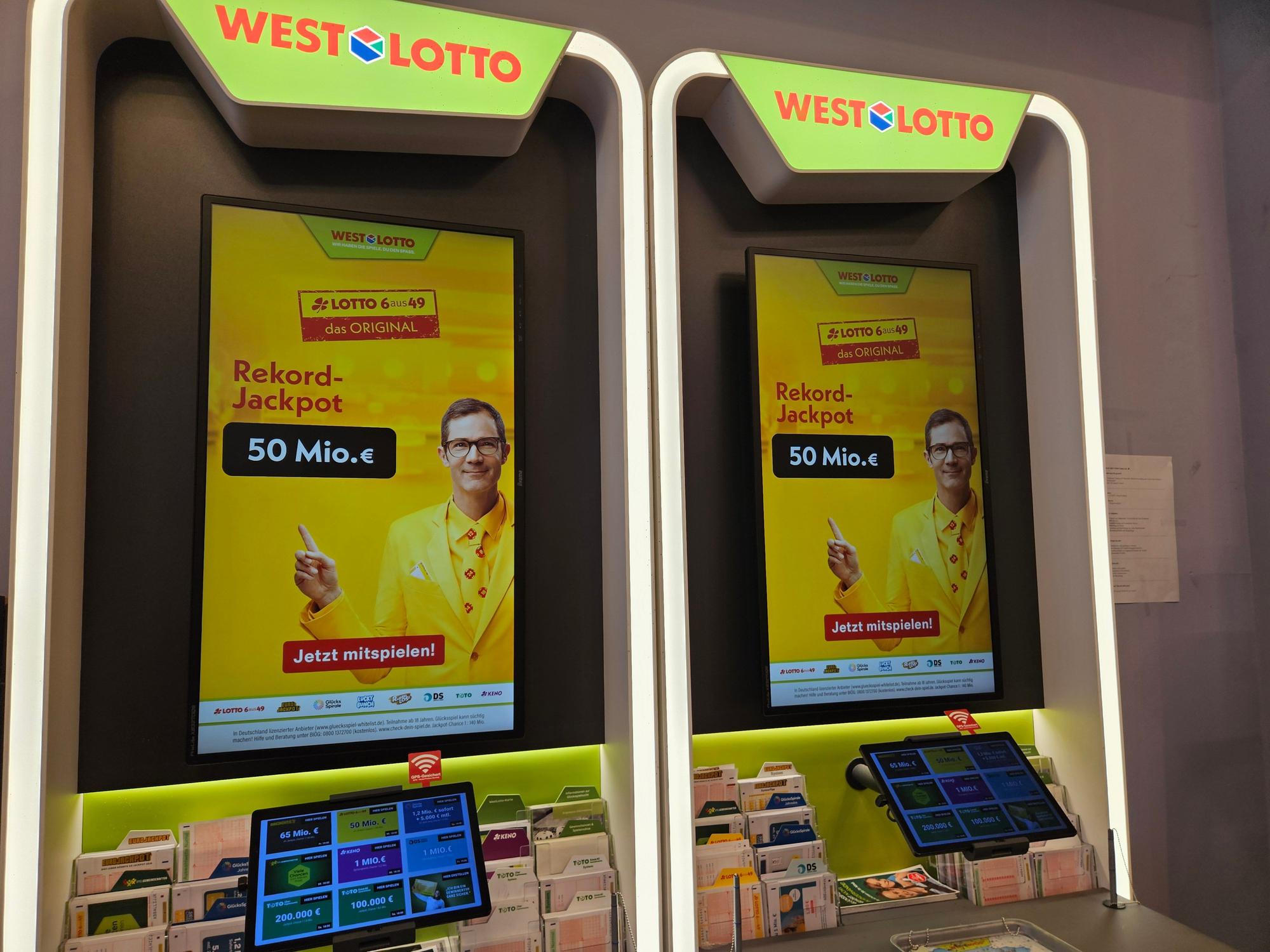 Auf dem Bild sieht man zwei WESTLOTTO-Terminals in einer Lotto-Annahmestelle mit großen Werbedisplays. Auf den Bildschirmen wird ein Rekord-Jackpot von 50 Millionen Euro für LOTTO 6aus49 beworben.