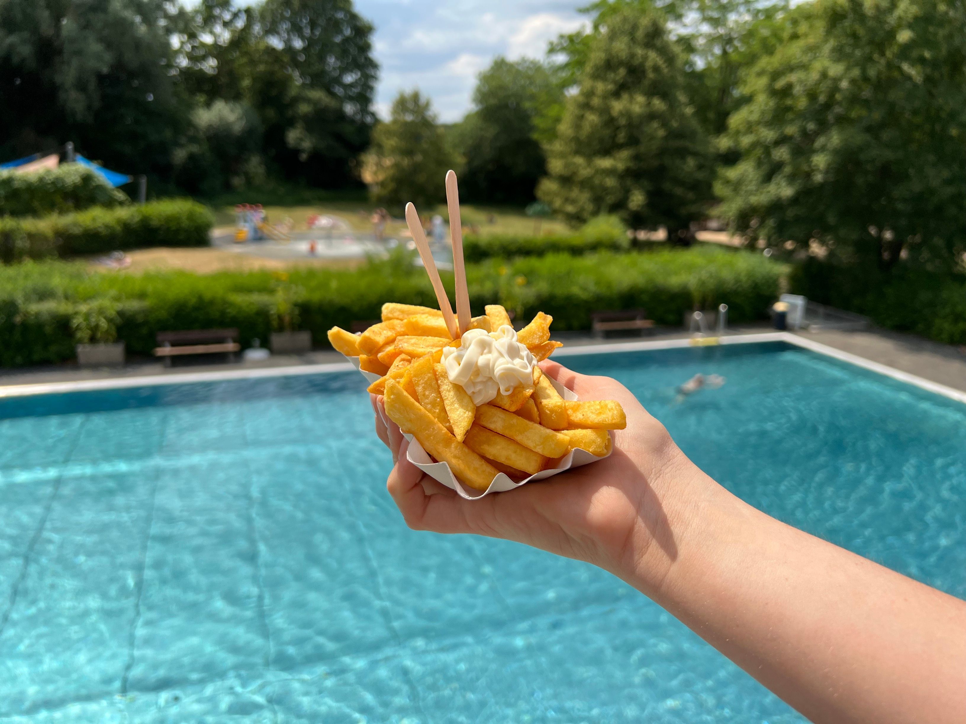 Eine Person hält in einem Freibad eine Portion Pommes vor die Kamera.